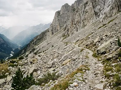 KI-generiertes Symbolbild: Schmaler Pfad über ein Geröllfeld unterhalb heller Felswände, tief darunter ein Tal mit Fluss