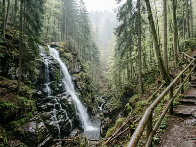 KI-generiertes Symbolbild: Wasserfall über moosige Felsstufen in einer bewaldeten Schlucht, rechts ein Treppenweg mit Holzgeländer